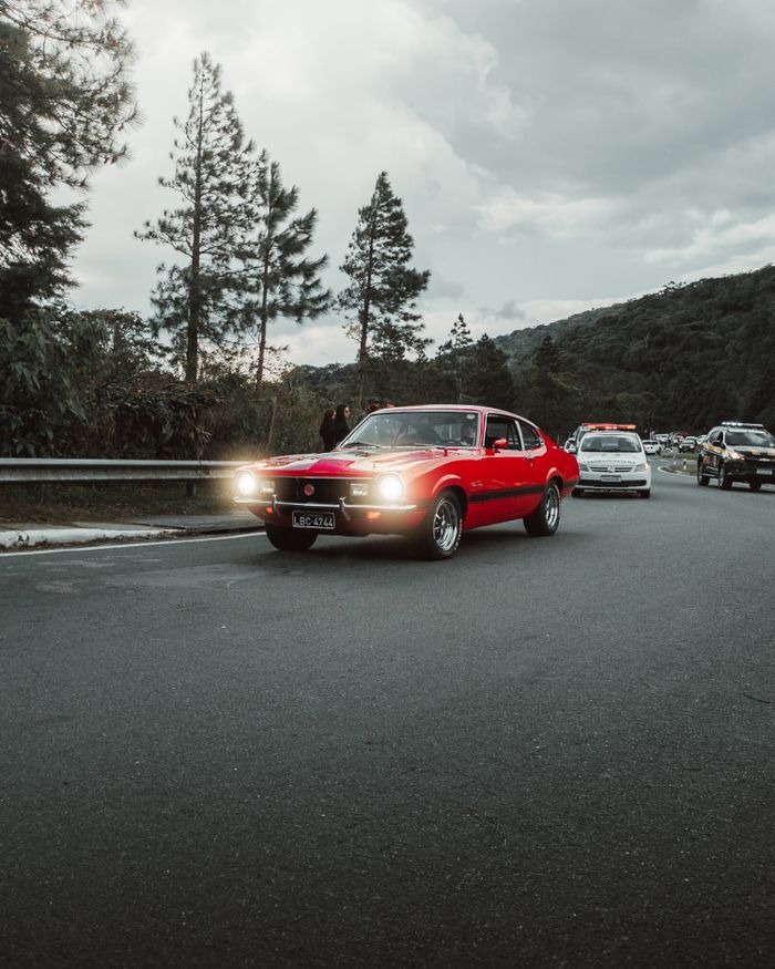 A classic red car driving on a scenic mountain road under a cloudy sky.