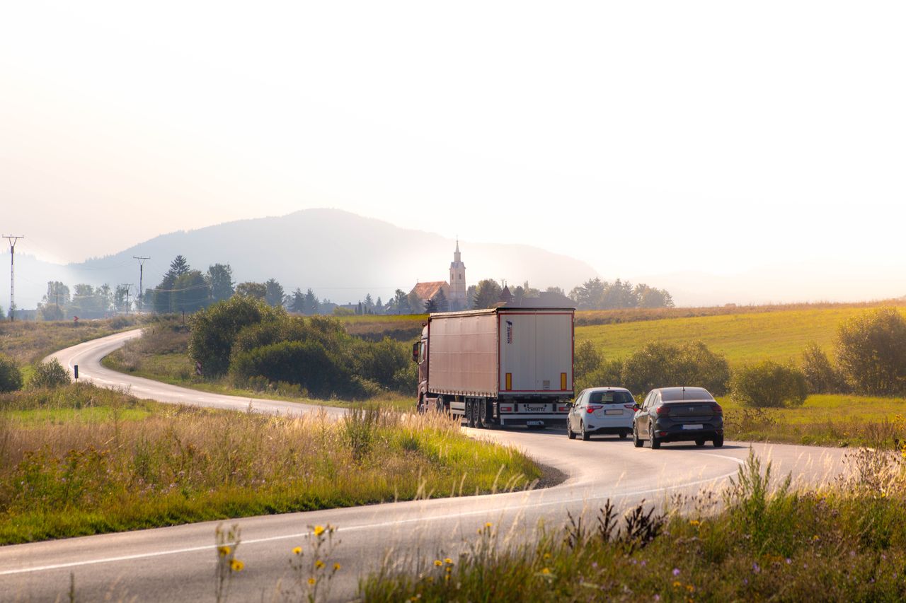Vehicles on a winding rural road with scenic mountain and field views during the day.