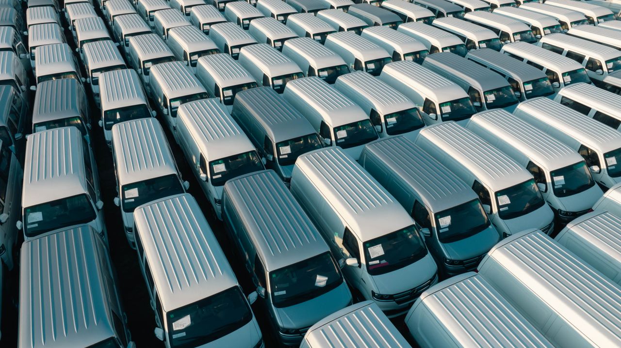 Aerial shot of multiple white vans parked in orderly rows in a lot under daylight.
