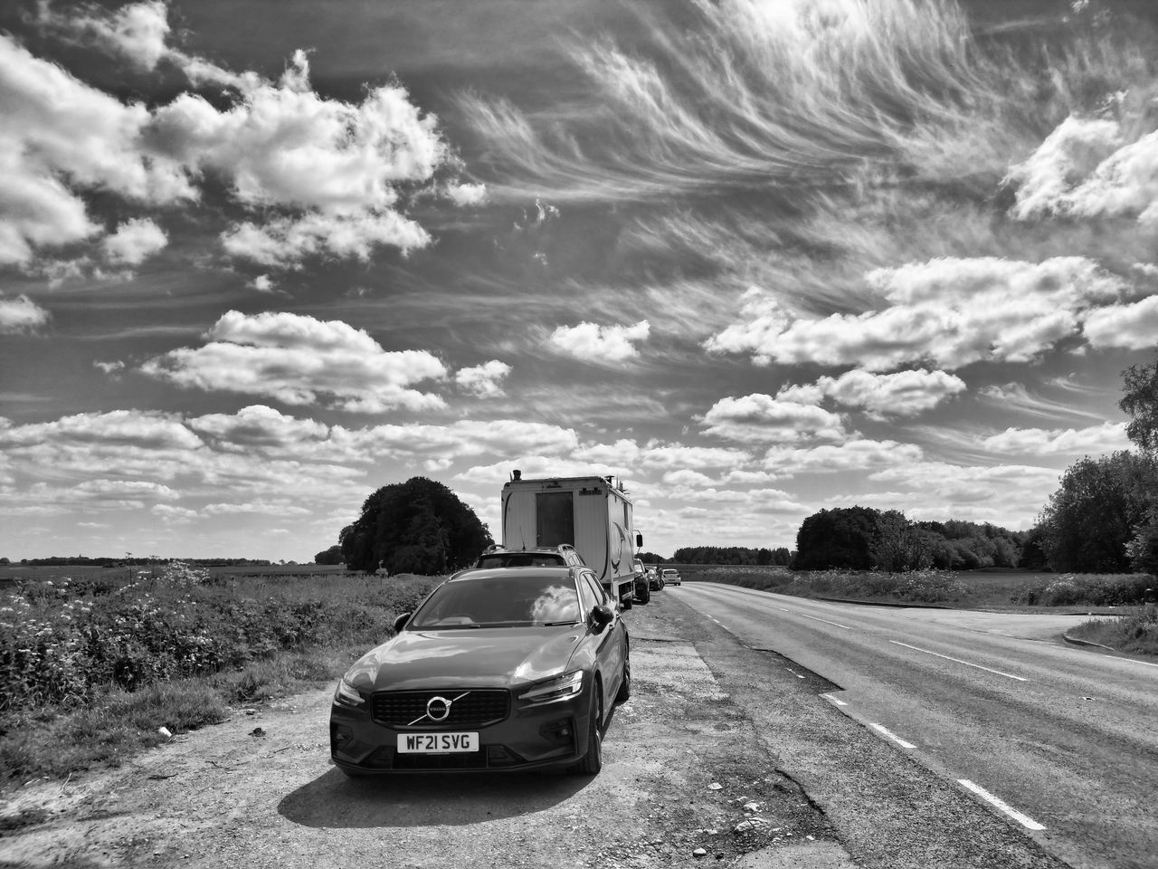 Monochrome view of a countryside road, with cars parked under a dynamic sky.